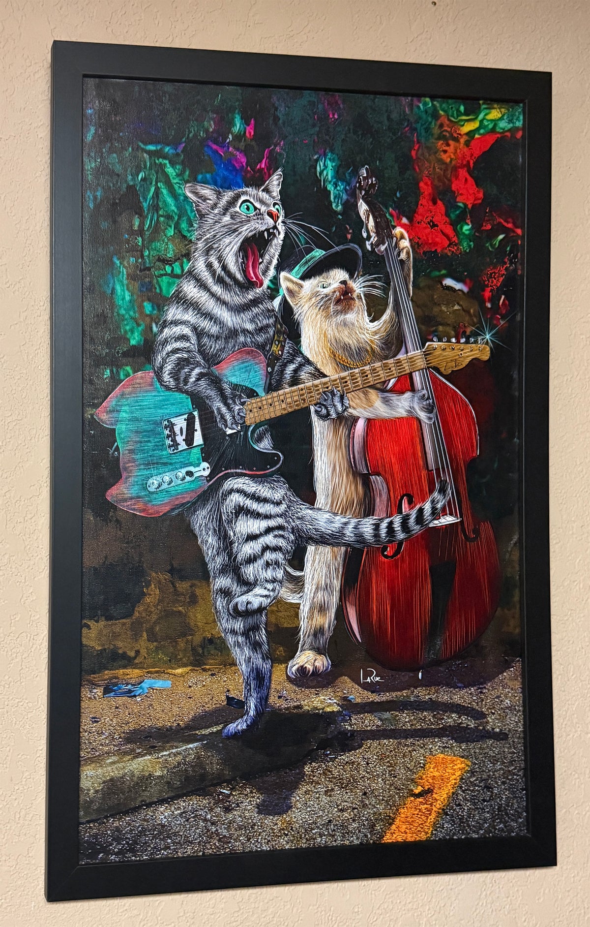Framed wall art featuring a surreal painting of two cats playing music, with one cat strumming a turquoise electric guitar and the other playing a red double bass, shown hanging on a beige wall. Buskers and Scruffy by Doug LaRue