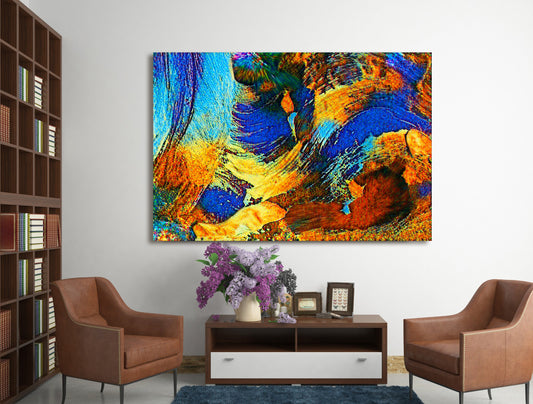 A large square abstract canvas print with vibrant blue, orange, yellow, and brown brushstrokes, displayed on a white wall in a modern living room setting with two brown armchairs and a dark wood console table. Rollingwood Cobalt by Doug LaRue