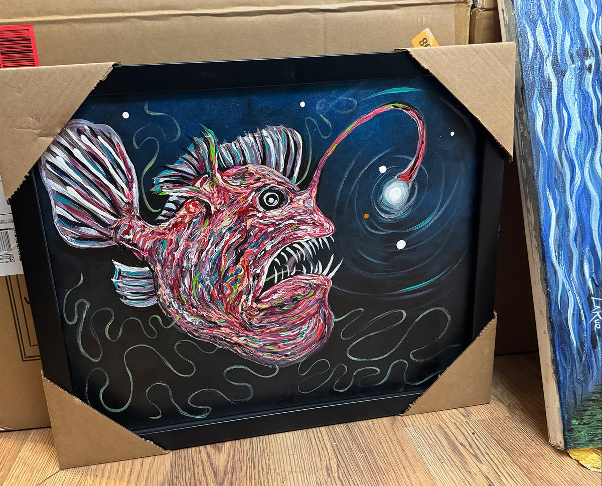 A canvas print featuring an original deep sea fish illustration, framed in a black museum style frame, displayed against a backdrop of packing materials.