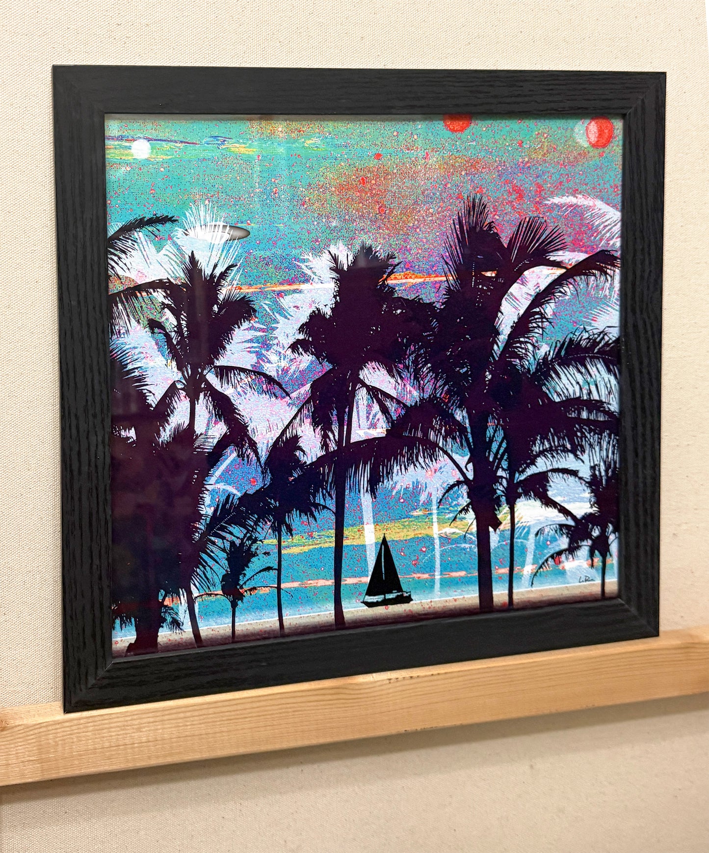 A framed square art print depicting a tropical beach scene with dark palm tree silhouettes, turquoise water, and a colorful sky, mounted on a light wood shelf against a beige wall.