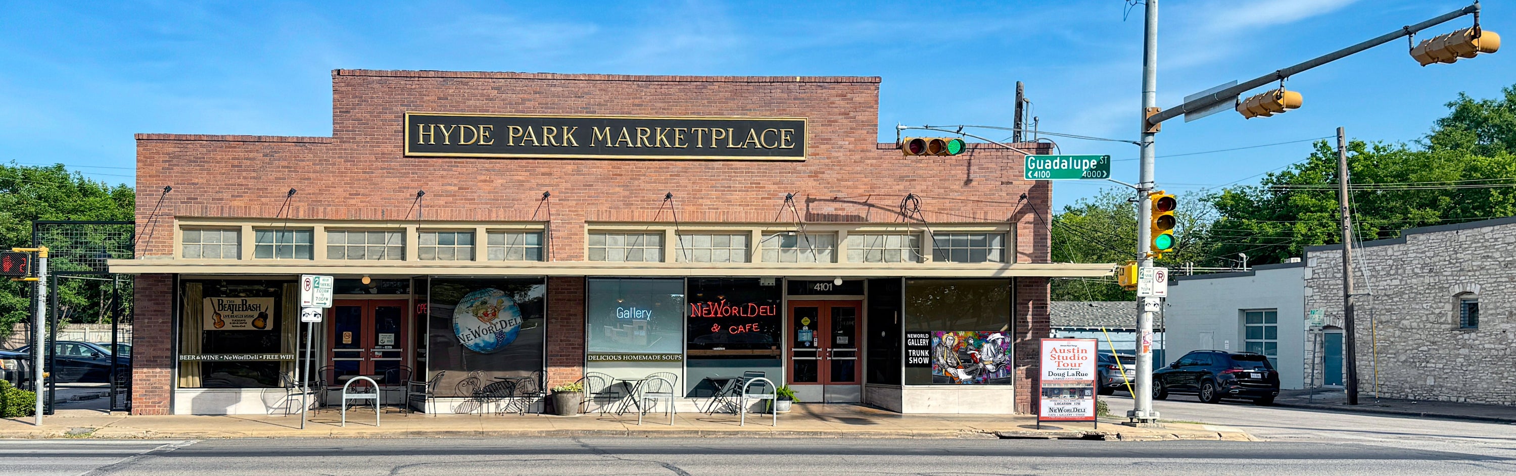 A brick storefront building labeled 'Hyde Park Marketplace' on the front, situated at a street intersection with traffic lights, trees, and a blue sky in the background.