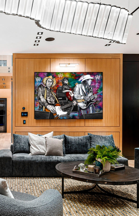A framed colorful wall art piece depicting three musicians playing instruments, displayed in a modern living room setting above a dark gray sofa and a round wooden coffee table. Slim, Red and Stevie Ray by Doug LaRue