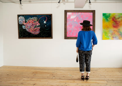 A person wearing a black hat, bright blue sweatshirt, and patterned pants stands with their back to the camera, viewing framed abstract paintings on a white gallery wall. Two framed artworks are visible, including one with a colorful fish and another with pink and red abstract patterns, set in a gallery with a light wood floor.
