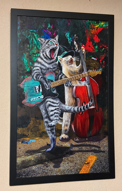 Framed wall art featuring a surreal painting of two cats playing music, with one cat strumming a turquoise electric guitar and the other playing a red double bass, shown hanging on a beige wall. Buskers and Scruffy by Doug LaRue