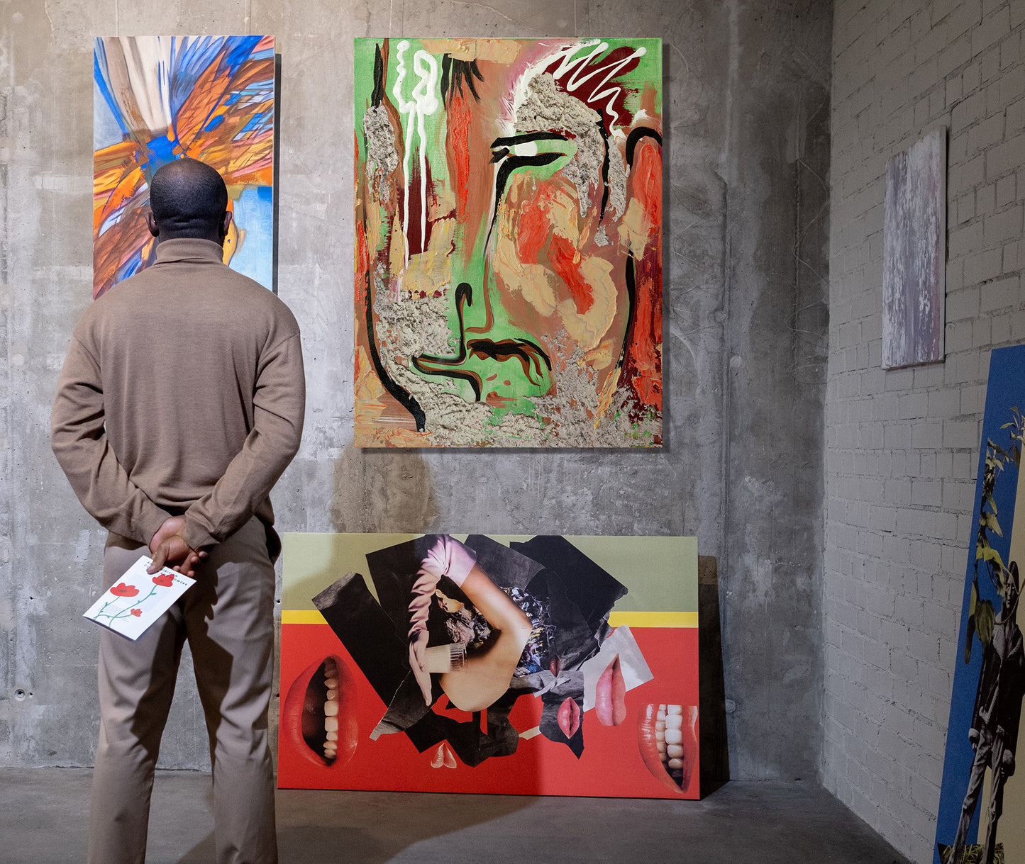A man in a beige tracksuit stands with his back to the camera, looking at a large abstract painting hanging on a gray concrete gallery wall. The artwork features a stylized face with green, red, black, and beige tones. Chuy by Doug LaRue