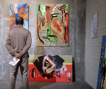 A man in a beige tracksuit stands with his back to the camera, looking at a large abstract painting hanging on a gray concrete gallery wall. The artwork features a stylized face with green, red, black, and beige tones. Chuy by Doug LaRue