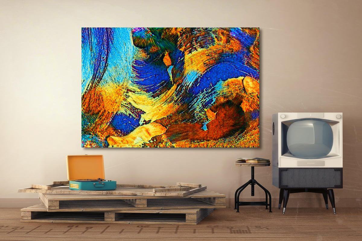 A large square abstract canvas print with vibrant blue, orange, yellow, and brown brushstrokes, displayed on a beige wall in a styled room setting with a wooden pallet coffee table, a vintage television, and a small stool. Rollingwood Cobalt by Doug LaRue