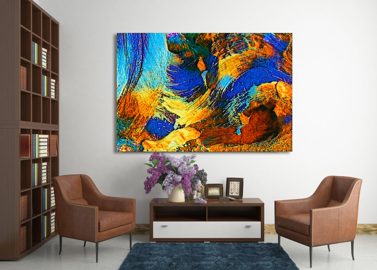 A large square abstract canvas print with vibrant blue, orange, yellow, and brown brushstrokes, displayed on a white wall in a modern living room setting with two brown armchairs and a dark wood console table. Rollingwood Cobalt by Doug LaRue