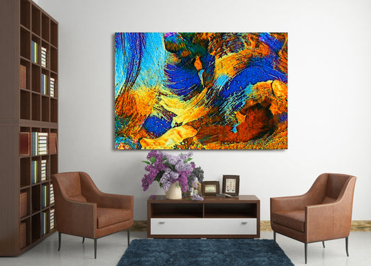 A large square abstract canvas print with vibrant blue, orange, yellow, and brown brushstrokes, displayed on a white wall in a modern living room setting with two brown armchairs and a dark wood console table. Rollingwood Cobalt by Doug LaRue