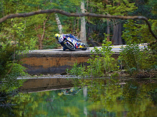 A motorcycle racer in a blue, white, and yellow racing suit leans heavily into a turn on a motorcycle on a dirt track, with a forest and water reflection in the background. Creek Bridge GP Motorcycle by Doug LaRue