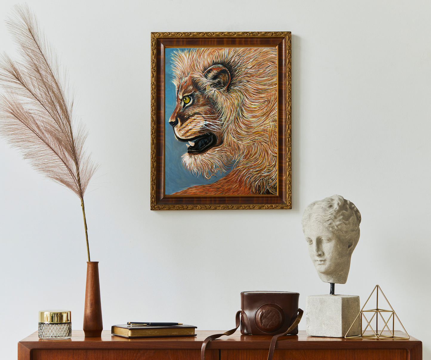 A framed painting of a lion's head in profile hangs on a light gray wall above a wooden console table styled with a vase of pampas grass, books, a stone bust, and a leather camera bag. Golden Lion oil painting by Doug LaRue