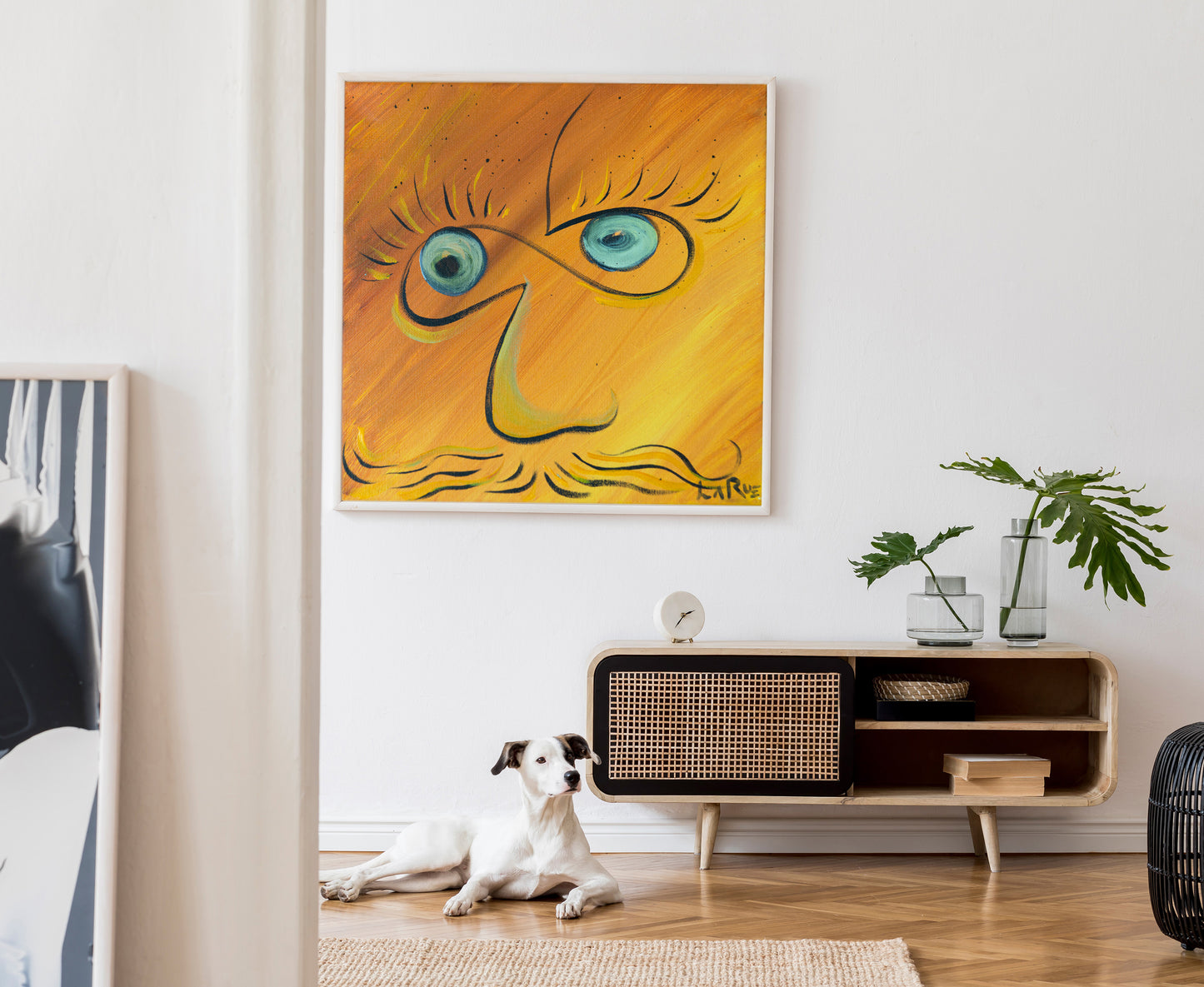 A framed abstract painting with a stylized face in yellow, orange, and blue hangs on a white wall above a wooden sideboard. A white dog lies on a woven rug on the floor below, and a potted plant sits on the cabinet.  Infinite Eyes #1 by Doug LaRue