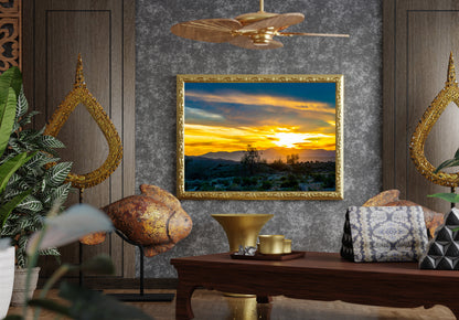 A framed landscape painting of a desert sunset is displayed on a textured gray wall in a styled room setting with a wooden table, decorative pillows, plants, and gold-toned wall mirrors.