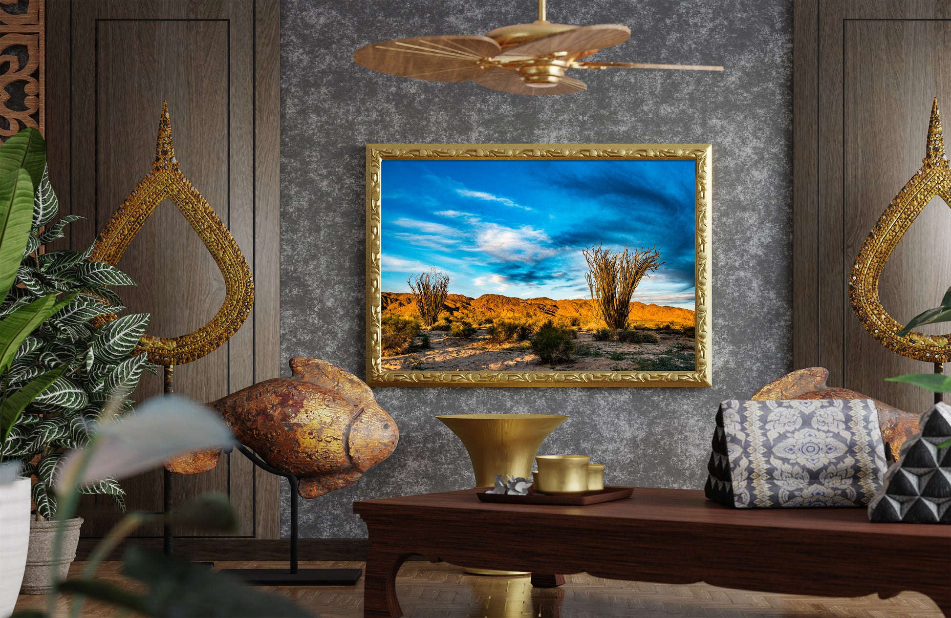 A framed landscape painting of a desert scene with cacti under a blue sky, displayed on a textured gray wall in a styled room with wooden furniture, plants, and decorative gold accents.