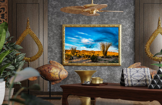A framed landscape painting of a desert scene with cacti under a blue sky, displayed on a textured gray wall in a styled room with wooden furniture, plants, and decorative gold accents.