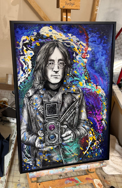 A framed portrait painting of John Lennon, depicted holding a camera, set against a vibrant abstract background with splashes of blue, purple, yellow, and white. The artwork is housed in a black frame and displayed on an easel in a studio or workshop environment.