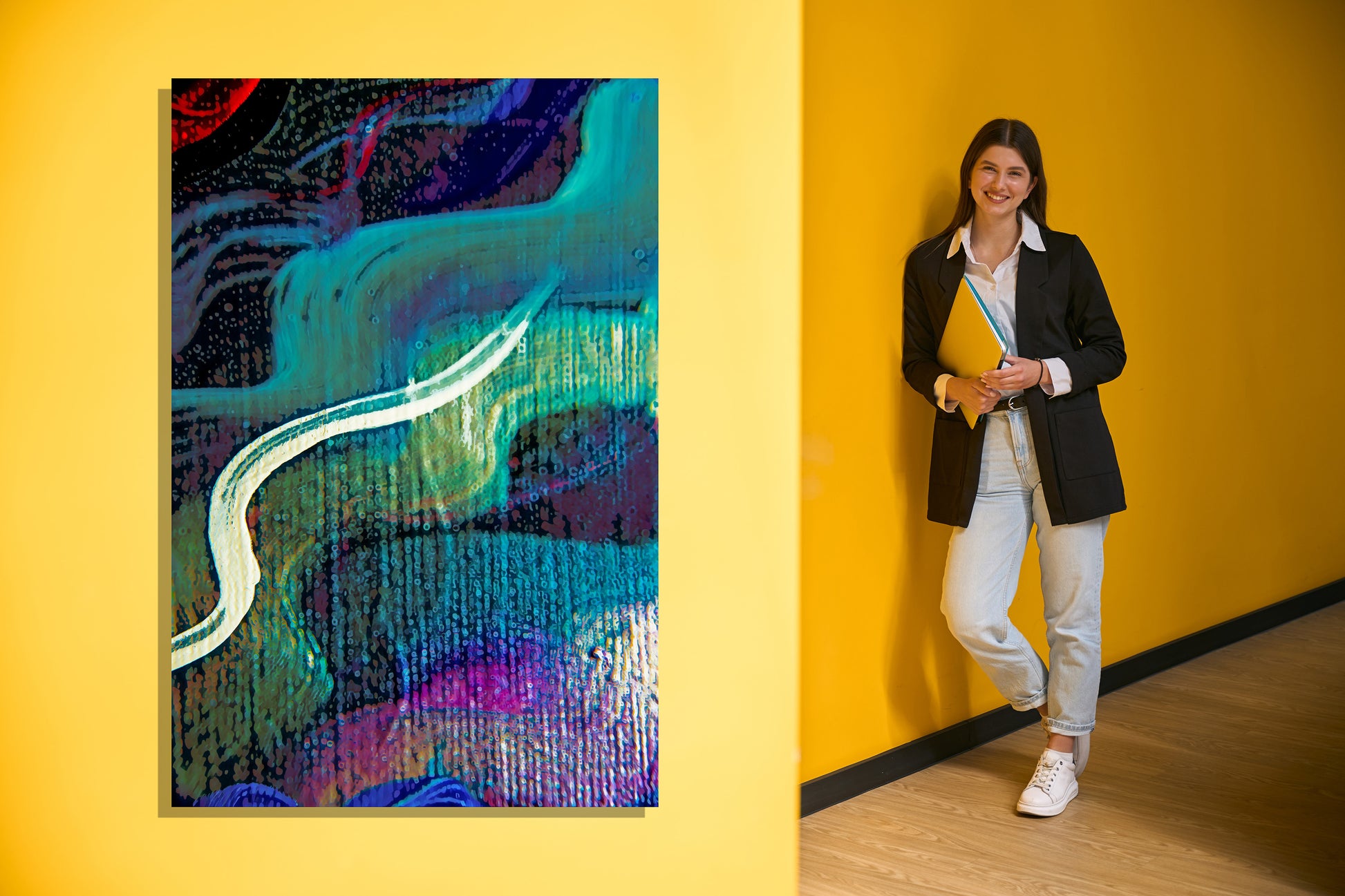 A woman in a black blazer, white shirt, and light jeans stands against a bright yellow wall holding a yellow folder, next to a colorful abstract art on the same yellow wall.  Lost Creek Feather art by Doug LaRue