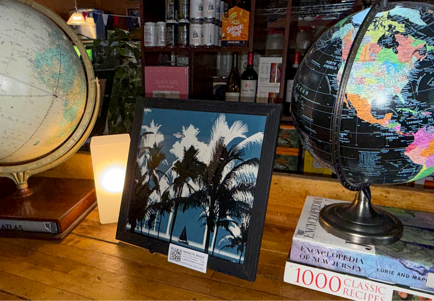 A framed art print depicting a tropical beach scene with palm trees under a starry night sky, styled on a wooden table next to two globe globes and books. Majestic Beach by Doug LaRue
