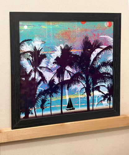 A framed square art print depicting a tropical beach scene with dark palm tree silhouettes, turquoise water, and a colorful sky, mounted on a light wood shelf against a beige wall.