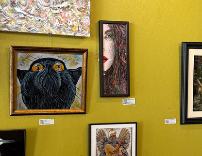 Artistic wall display with various framed artworks on a yellow wall. TAra's Green eye 19x9 art print in a black wood frame.